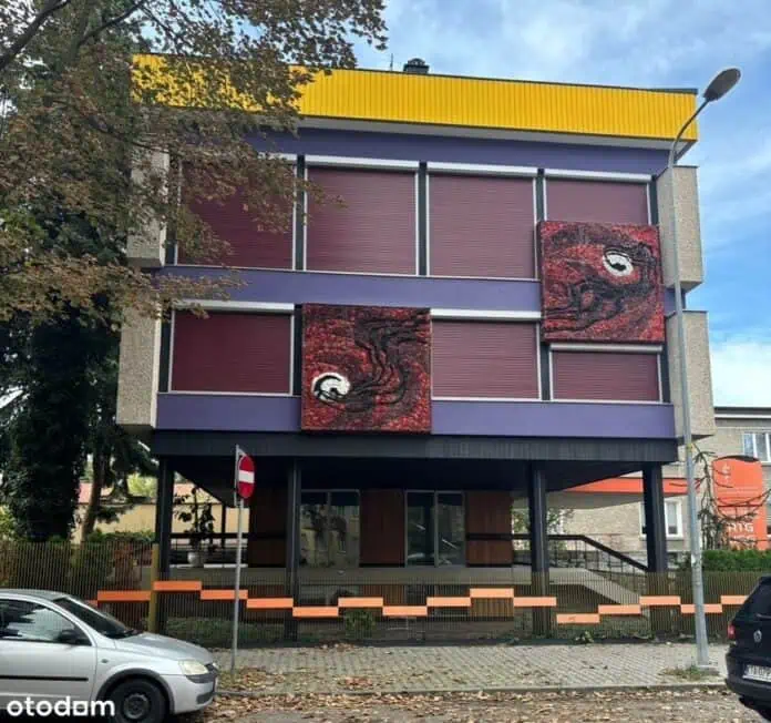 Zewnętrzna fasada budynku z fioletowymi panelami, czerwonymi mozaikami i żółtym gzymsem.